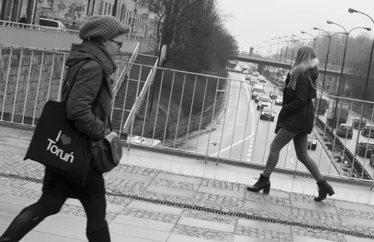 Woman with an I Love Torun tote passing a blonde woman on a pedestrian bridge above traffic